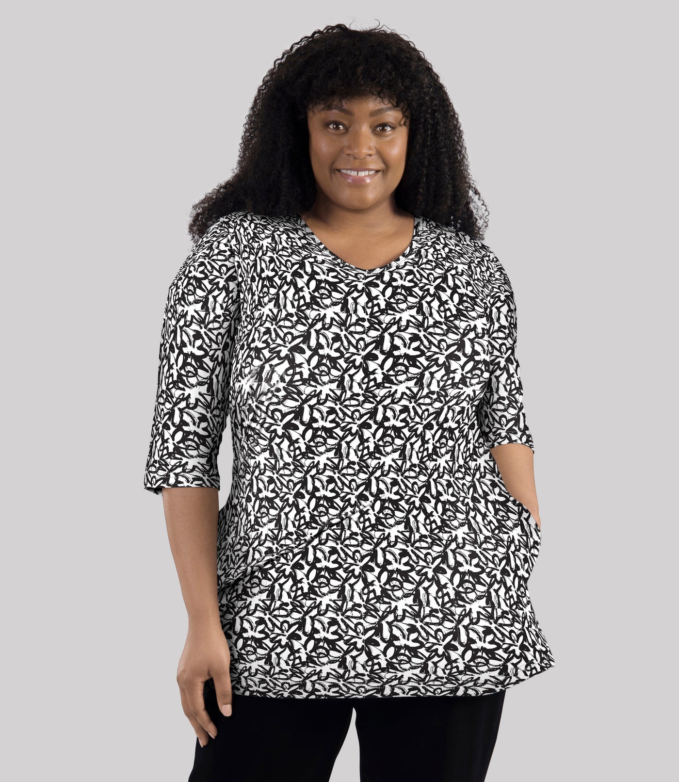 Model wearing JunoActive's Junonia Lifestyle Cotton Printed 3/4 sleeve pocket tunic in pattern black and white wildflower. Her left hand is in left pocket of shirt.