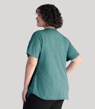 Model, facing back, wearing Softwik scoop neck short sleeve plus size top in heather emerald. Model is wearing black pants.