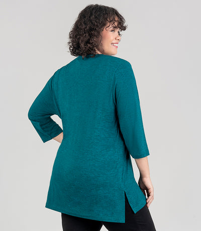 JunoActive model facing back, wearing SoftSupreme V Neck 3-4 sleeve tunic in color dark heather teal. Her right arm is by her side and left arm is bent on her hip. Tunic is longer in back with a side slit.