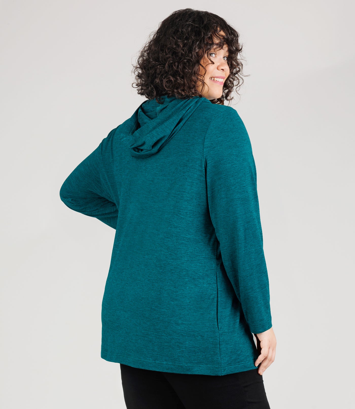 JunoActive model, facing back, wearing SoftSupreme Pocketed Hoodie in color Heather Dark Teal. Model's left hand is on left hip and right arm dropped by her side.