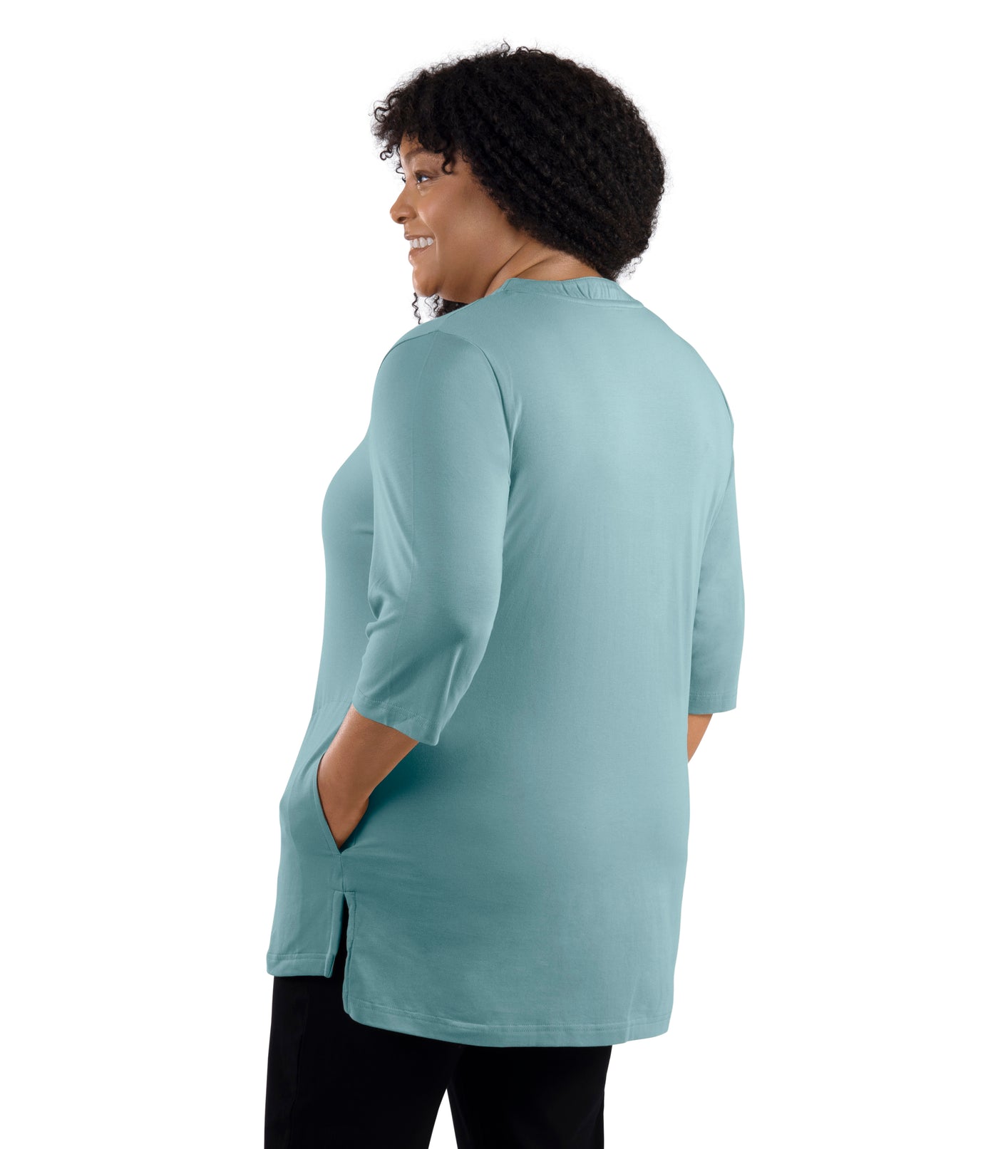 JunoActive Model wearing Stretch Naturals Lite 3/4 Sleeve Button Henley, facing back, one hand in pocket of shirt and other by her side. Color calm jade.