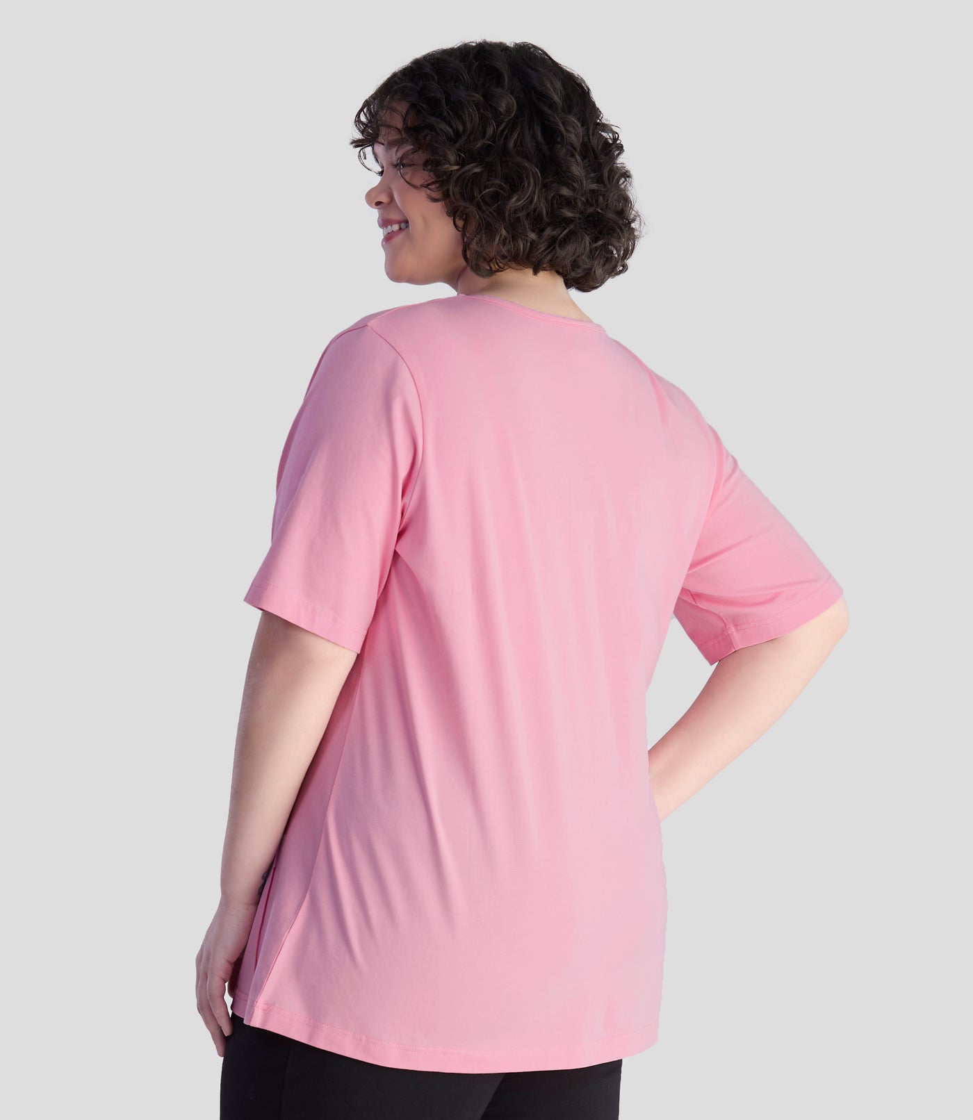 JunoActive model, wearing Stretch Naturals Lite Pintuck Top in color peony pink. Model is facing back, left arm is draping by her side and right hand is on her right hip.
