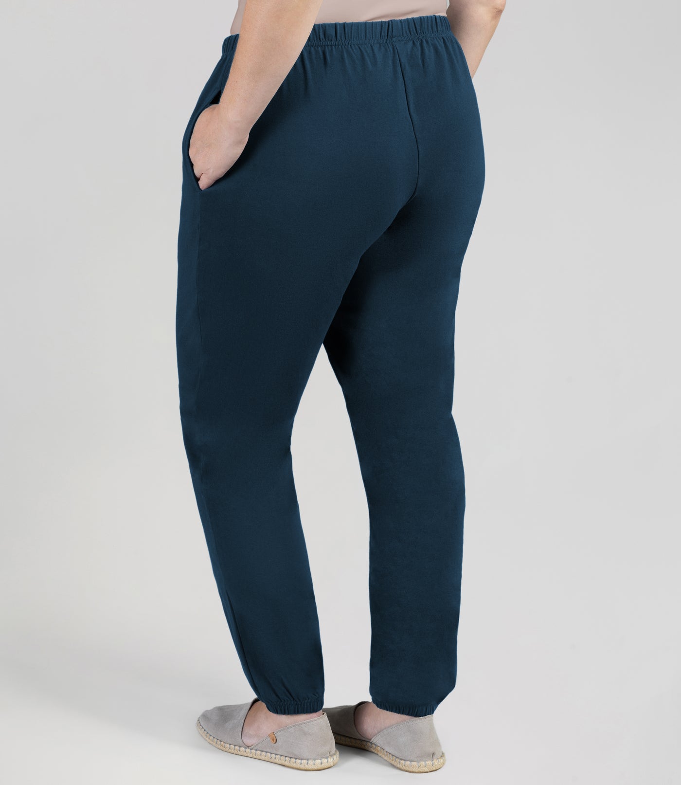 Plus size woman facing back is wearing JunoActive Stretch Naturals plus size jogger pant bottom. Her left hand is in pocket and right arm hanging by side. The pant is shirred at top with elastic band and loosely fitted. The hem at bottom is gathered with elastic at ankle. Color is indigo.