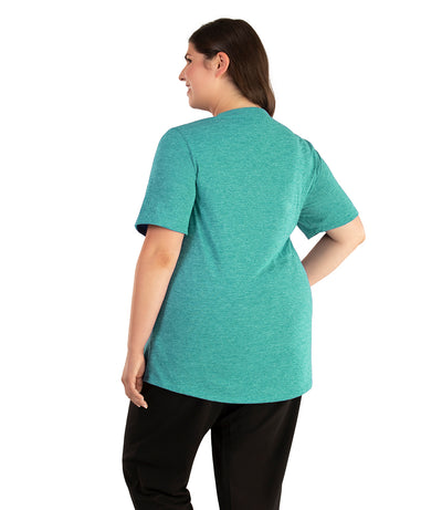 Plus size woman, facing back looking to the left, wearing JunoActive plus size QuikLite Scoop Neck Short Sleeve top in the color Fern Green. She is wearing JunoActive Plus Size Leggings in the color black. 