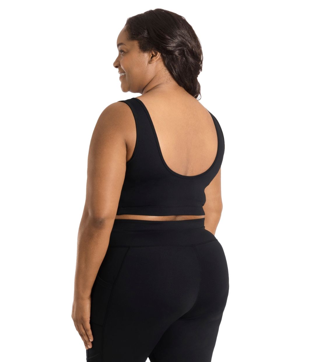 Plus size woman, facing back, wearing JunoActive plus size JunoStretch Scoop Bra in black. The woman is wearing black plus size JunoActive leggings. Her arms fall naturally to her side with her hands on her thighs.