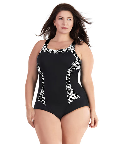 Plus size woman, facing front, wearing JunoActive plus size QuikEnergy Color Block Tank Suit Black and Hibiscus print. The blocking at the neckline and princess panels is Hibiscus print. Side panels, straps, center front, and leg blocking are black, has a scoop neckline and conservative leg opening.