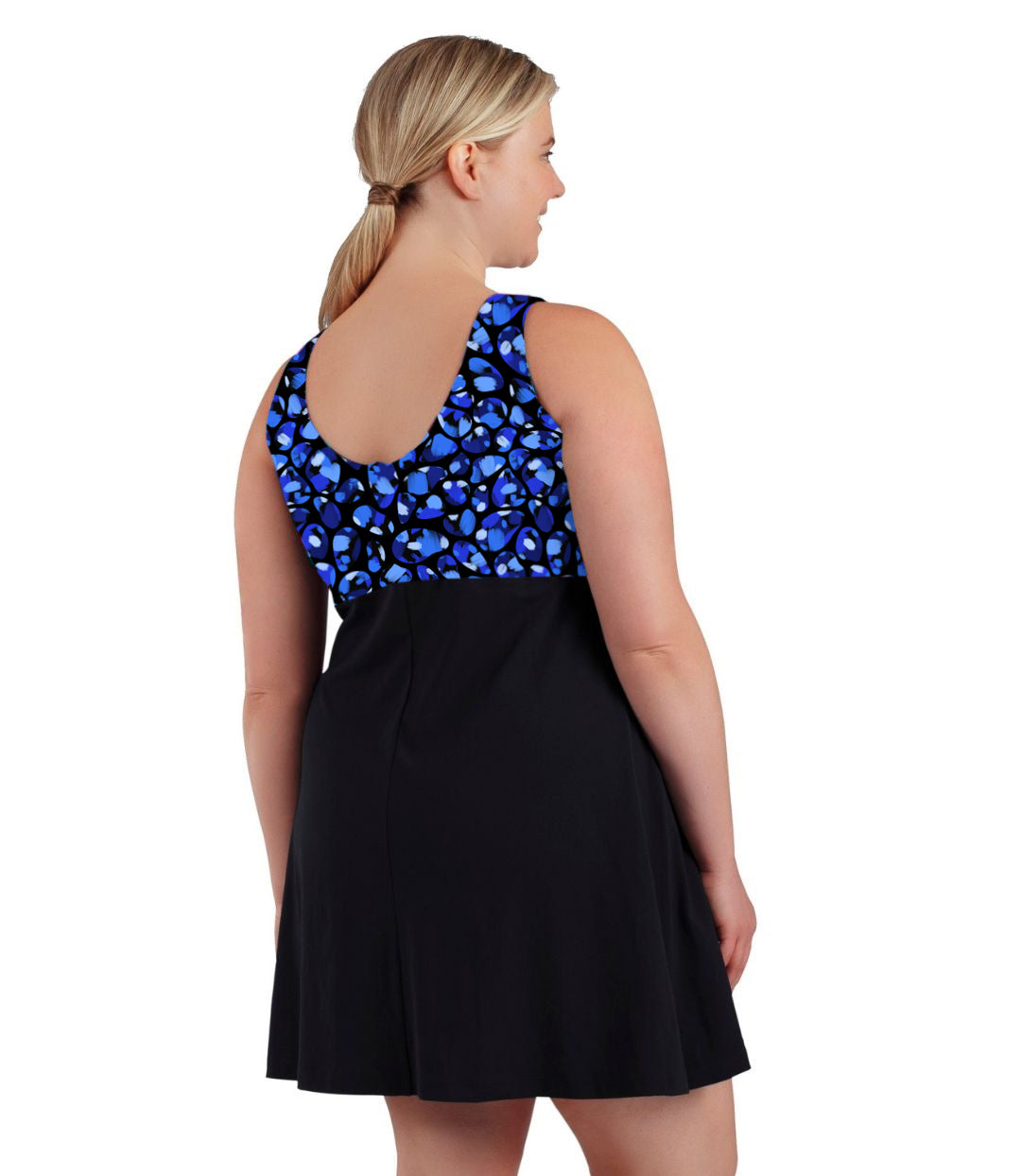 Plus size woman, back view, wearing JunoActive plus size Aquasport Zip Front Swim Dress Ocean Blues Print Black. The top of the swim dress has a multi colored blue bubble print and scoop back. The bottom is solid black, has a slight a-line shape and ends mid-thigh.