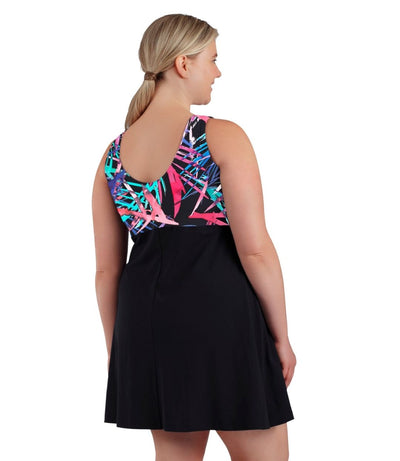 Plus size woman, back view, wearing JunoActive plus size Aquasport Zip Front Swim Dress Sunset Palm Print Black. The top of the swim dress has a multi colored pink blue and black Palm print and scoop back. The bottom is solid black, has a slight a-line shape and ends mid-thigh.
