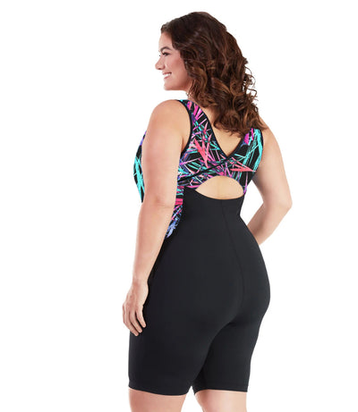 Plus size woman, back view, wearing JunoActive plus size AquaSport Crossback Aquatard Sunset Palm Print Black. The overlapping crossback and side blocking detail of the tanksuit is a multi colored pink blue and black Palm print. The main body of the suit is solid black, keyhole opening at the mid-back. The leg hits a few inches above the knee.