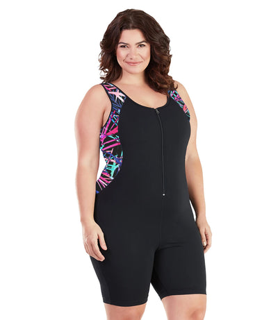 Plus size woman, facing front, wearing JunoActive plus size AquaSport Crossback Aquatard Sunset Palm Print Black. Front princess seam blocking is a multi colored pink blue and black Palm print.  The main body of the suit is solid black, has a black zipper in front with black bindind and the neck and armholes. The leg hits a few inches above the knee.
