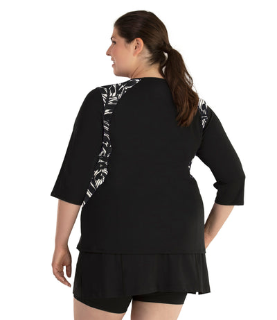 Plus size woman, facing back and side profile, wearing AquaSport Three Quarter Sleeve Rash Guard Tropical Print. V-neckline, black tropical print torso and black colorblocked sleeves and side of waist. She is wearing a pair of black JunoActive plus size swim shorts.