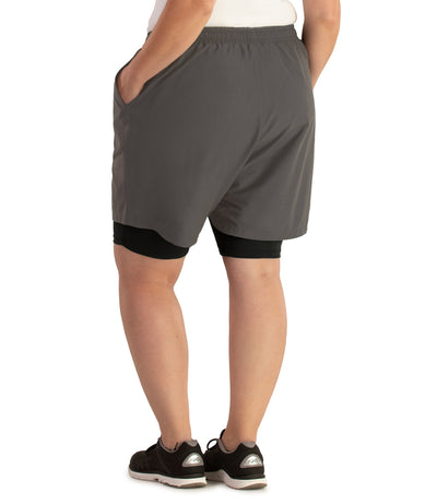 Bottom half of plus sized woman, facing back to viewer, hand in pocket, wearing JunoActive Dual Layer Walking Short in black and oak gray. These shorts have two layers with the bottom being a black tight bike short and the outer is shell is like material and looser fitted in oak gray. The hem is above knees.