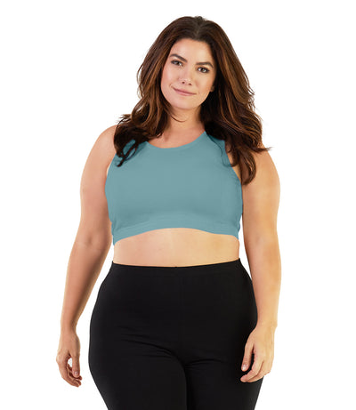 Plus size woman, facing front, wearing JunoActive plus size Stretch Naturals Scoop Neck Bra in soft green. The woman is wearing black JunoActive plus size leggings. Her arms fall naturally to her side. 