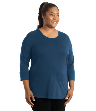 Plus size woman, facing front, wearing JunoActive’s Stretch Naturals Center Seam Scoop Neck 3/4 sleeve top in color denim blue. Hands are by side. Pants are in color black. 