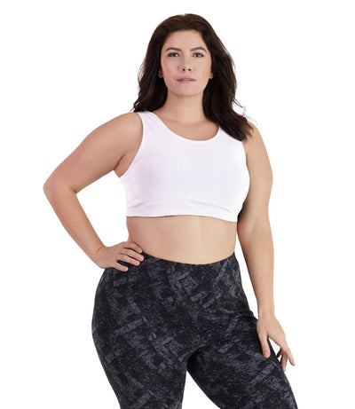 Plus size woman, facing front, wearing JunoActive plus size Stretch Naturals Scoop Neck Bra in white. The woman is wearing black JunoActive plus size leggings. Her right arm is on her hip, her left arm falls naturally to her side. 