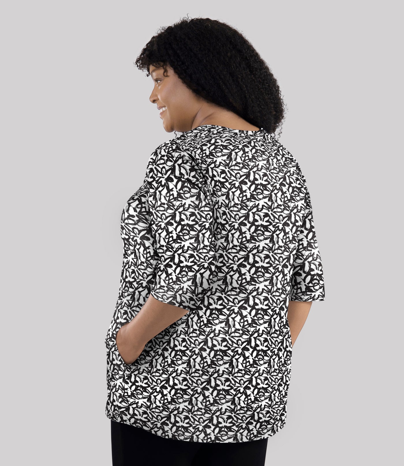 Model, facing back, wearing JunoActive's Junonia Lifestyle Cotton Printed 3/4 sleeve pocket tunic in pattern black and white wildflower. Her left hand is in left pocket of shirt.