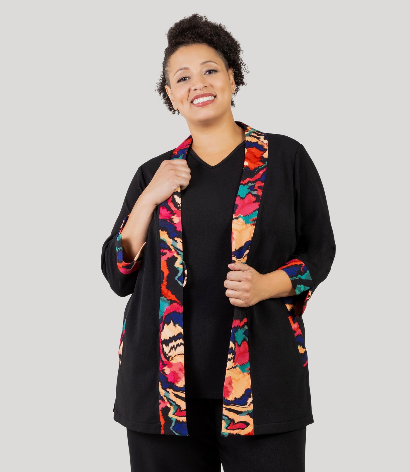 Plus size woman, facing front, wearing a JunoActive's Junonia Classic Plus size cardigan featuring modern ikat print on lapels and cuffs with remainder solid black. Cardigan is unbuttoned and her hands are holding onto lapels.
