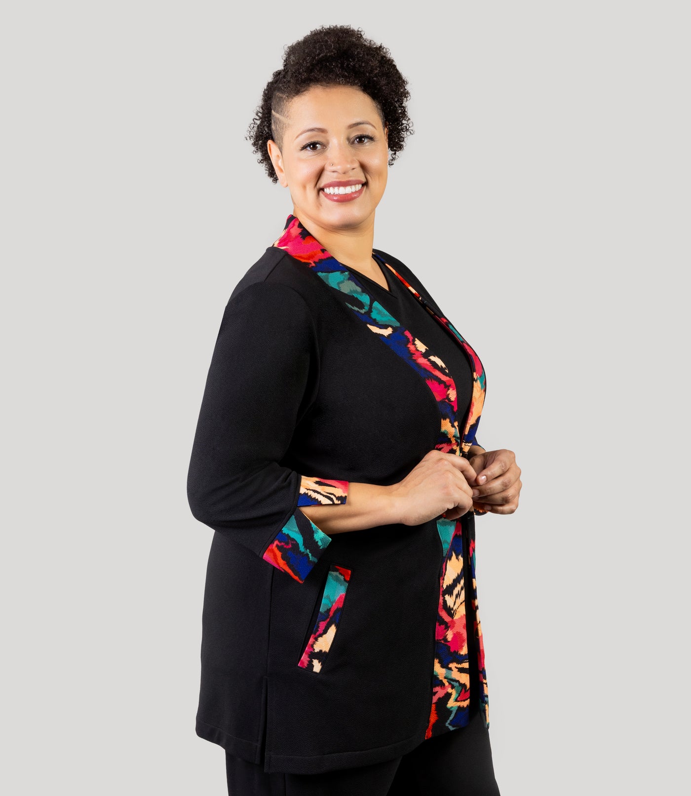 Plus size woman, facing front and side, wearing a JunoActive's Junonia Classic Plus size cardigan featuring modern ikat print on lapel and cuffs and solid black for the reminder of cardigan.