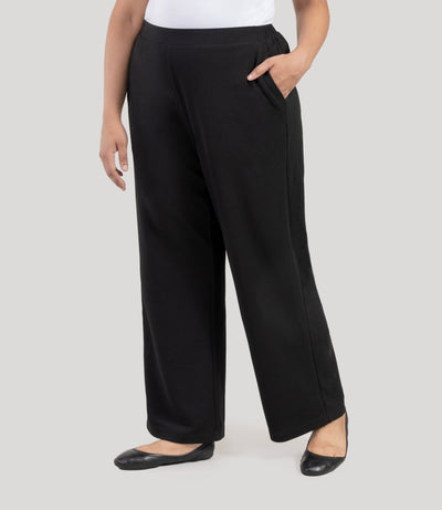 Plus size woman, facing side, wearing JunoActive's Junonia Classic Pocketed black Plus Size pants on a plain  background.