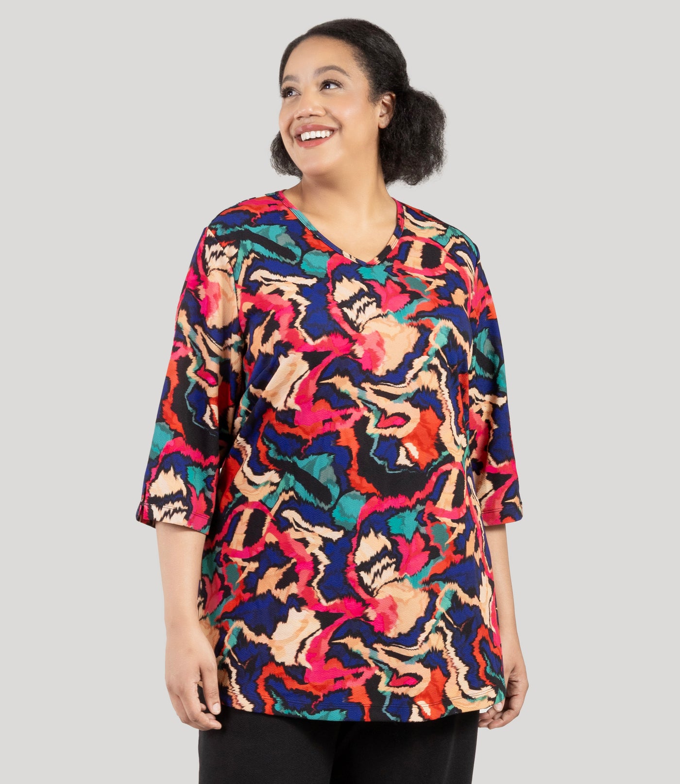 Plus size woman, facing front, wearing a JunoActive's Junonia Classic 3/4 Sleeve V-Neck Plus Size Tunic. Top features modern ikat print, v-neck, 3/4 sleeves and side vents at hem. Model iis looking to the side but body is facing straight on.