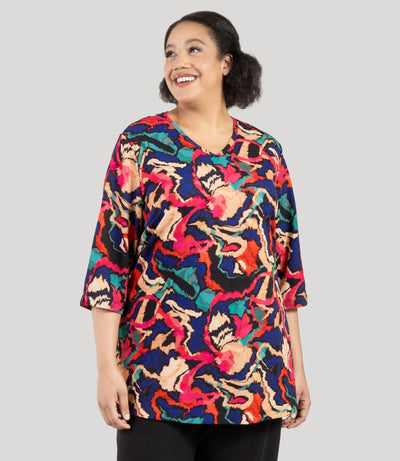 Plus size woman, facing front, wearing a JunoActive's Junonia Classic 3/4 Sleeve V-Neck Plus Size Tunic. Top features modern ikat print, v-neck, 3/4 sleeves and side vents at hem. Model iis looking to the side but body is facing straight on.