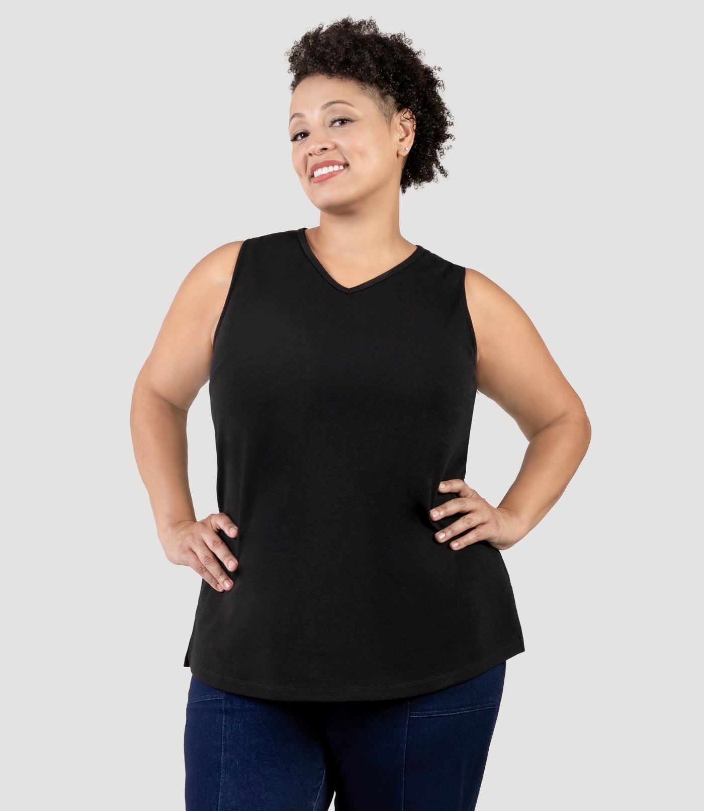 Plus size Woman, facing front, wearing JunoActive's Junonia Classic Sleeveless Plus Size Top in black,  against a plain background. Arms on her hips. Sleeveless top features v-neck and side hem vents.