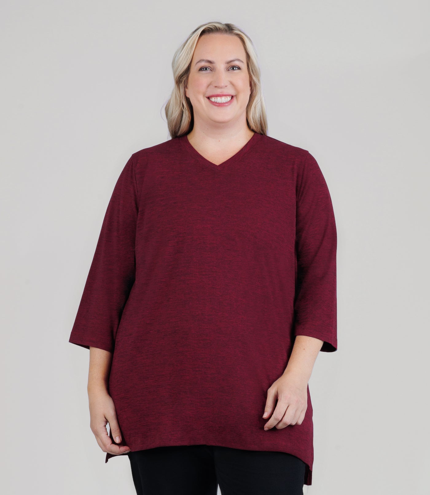 JunoActive model facing front, wearing SoftSupreme V Neck 3-4 sleeve tunic in color heather cranberry. Both of models arms and hands resting by her side. Tunic is longer in back with a side slit.