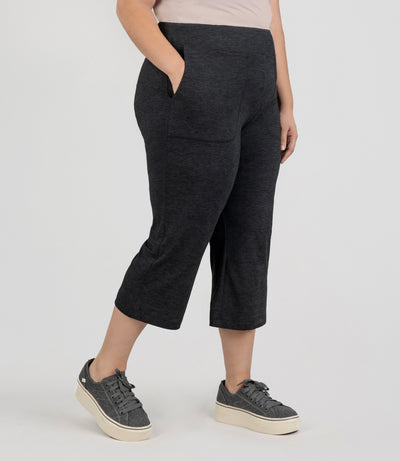 JunoActive model, facing side, wearing SoftSupreme Pocketed Lounge Capri in color heather black. Right hand in pocket of capri and left arm hanging by side.