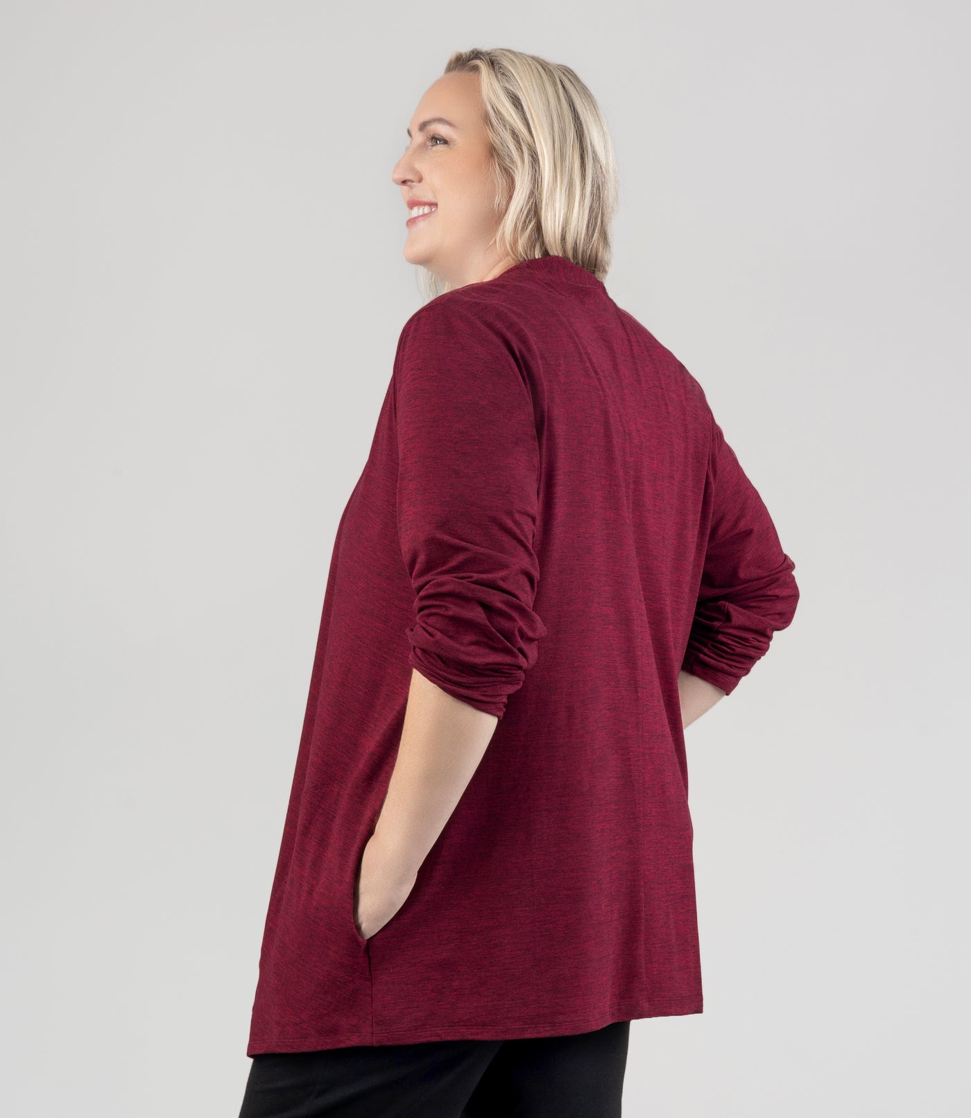 JunoActive Model, facing back, wearing SoftSupreme Cardigan in Heather Cranberry. Sleeves rolled up to elbows and hands in pockets of cardigan.