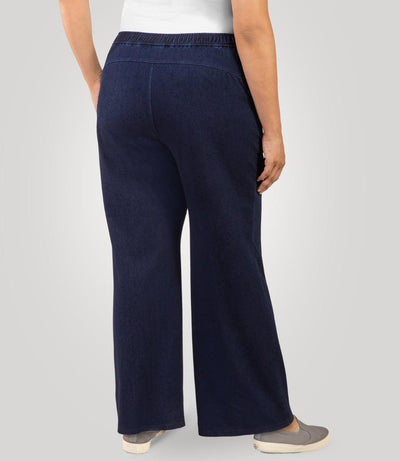 Plus size woman, facing back, wearing JunoActive's Wide Leg Stretch Twill Plus Size Pant. Her left hand is in her left pant pocket and right hand and arm is draping on her right side. Woman is standing on a beige colored backdrop.