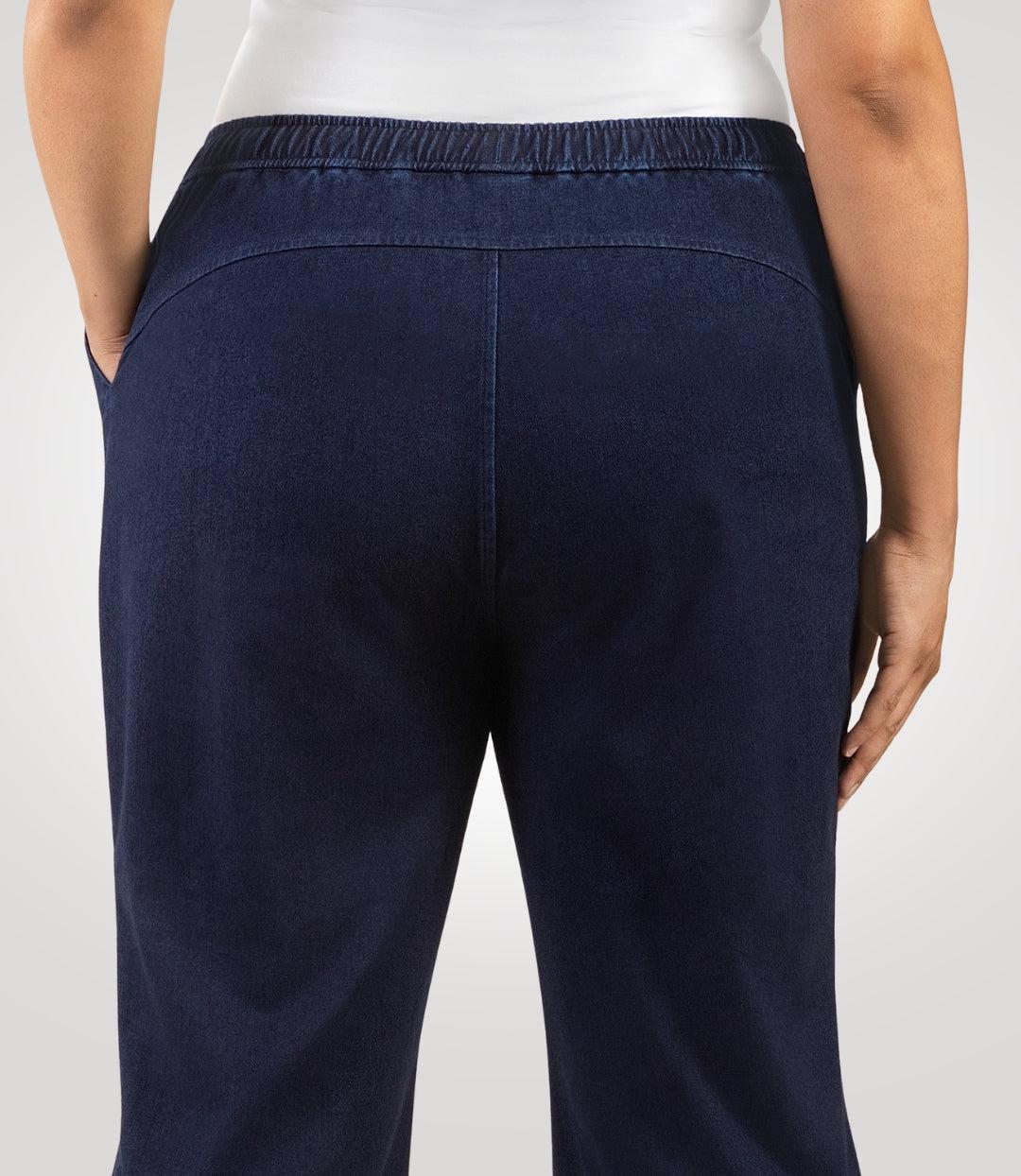 Plus size woman, facing back and close up of product, wearing JunoActive's Wide Leg Stretch Twill Plus Size Pant. Her left hand is in her left pant pocket and right hand and arm is draping on her right side. Woman is standing on a beige colored backdrop.