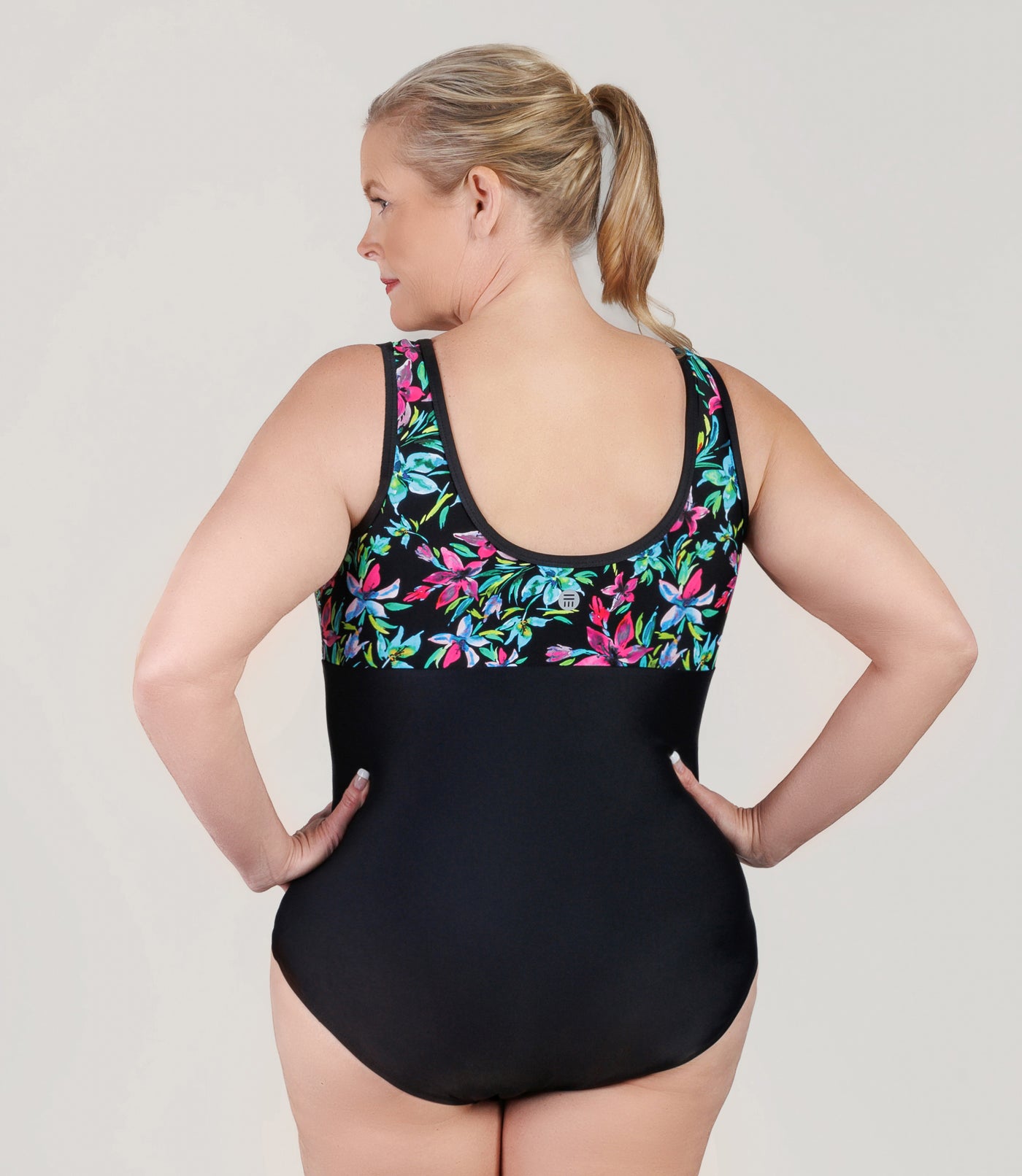 Plus size woman wearing JunoActive's QuikEnergy Empire plus size swimming suit in night Lillies print black. Models hands are on her hips and facing backwards.