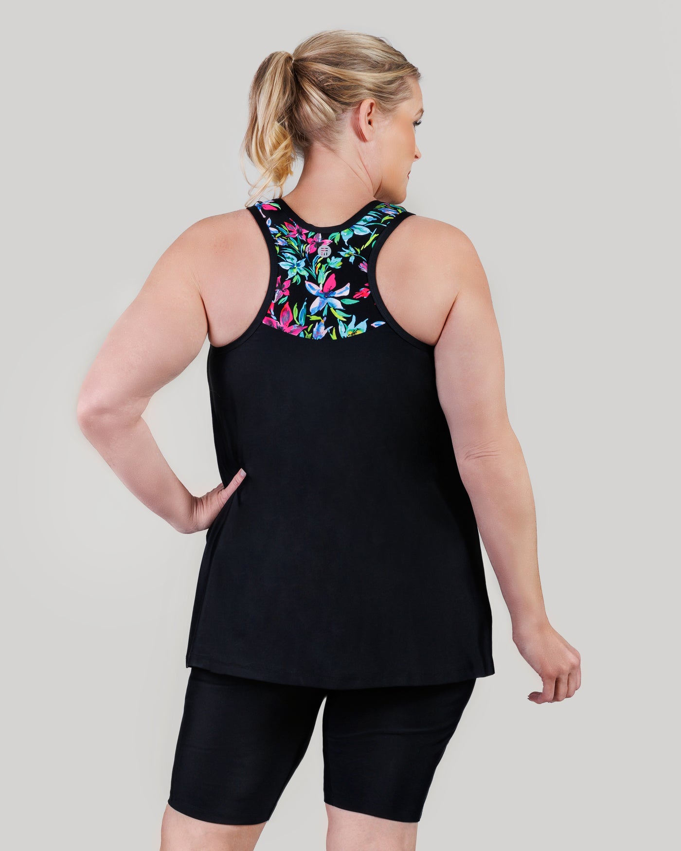 Plus size model, facing back, wearing JunoActive's QuikEnergy Kona plus size tankini, featuring Night Lillies and black print. Her left hand is on her left hip and right arm by her side.