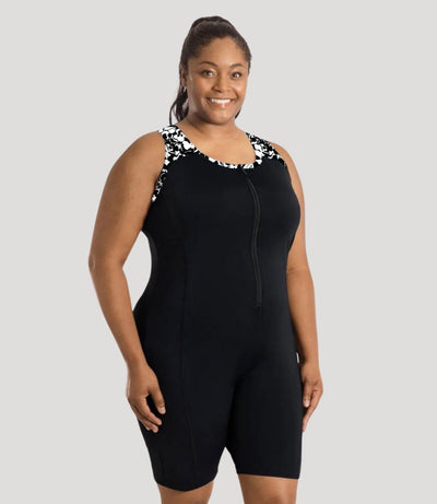 Plus size woman, facing front, wearing JunoActive plus size QuikEnergy Racerback Zip Front Aquatard Hibiscus print. Neck binding, armhole binding, and front shoulder blocking is a black and white print. The main body of the suit is solid black, has a black zipper in front. The leg hits a few inches above the knee.