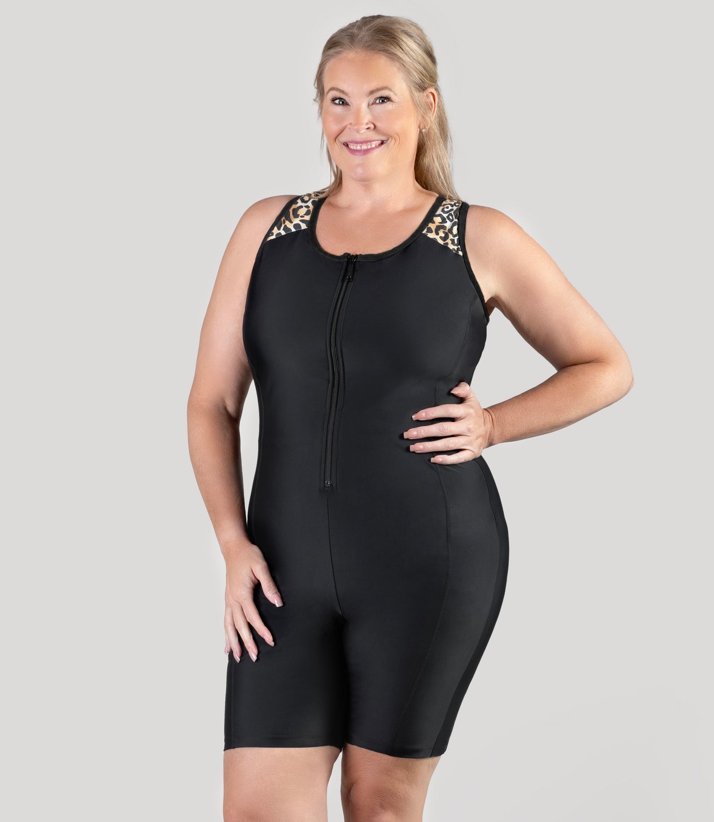 Plus size model, facing front, wearing JunoActive's QuikEnergy Racerback Zip Front plus size Aquatard. Left hand on hip and right hand is resting on leg. This plus size swimming suit features a fun leopard print on the shoulder straps and racerback.