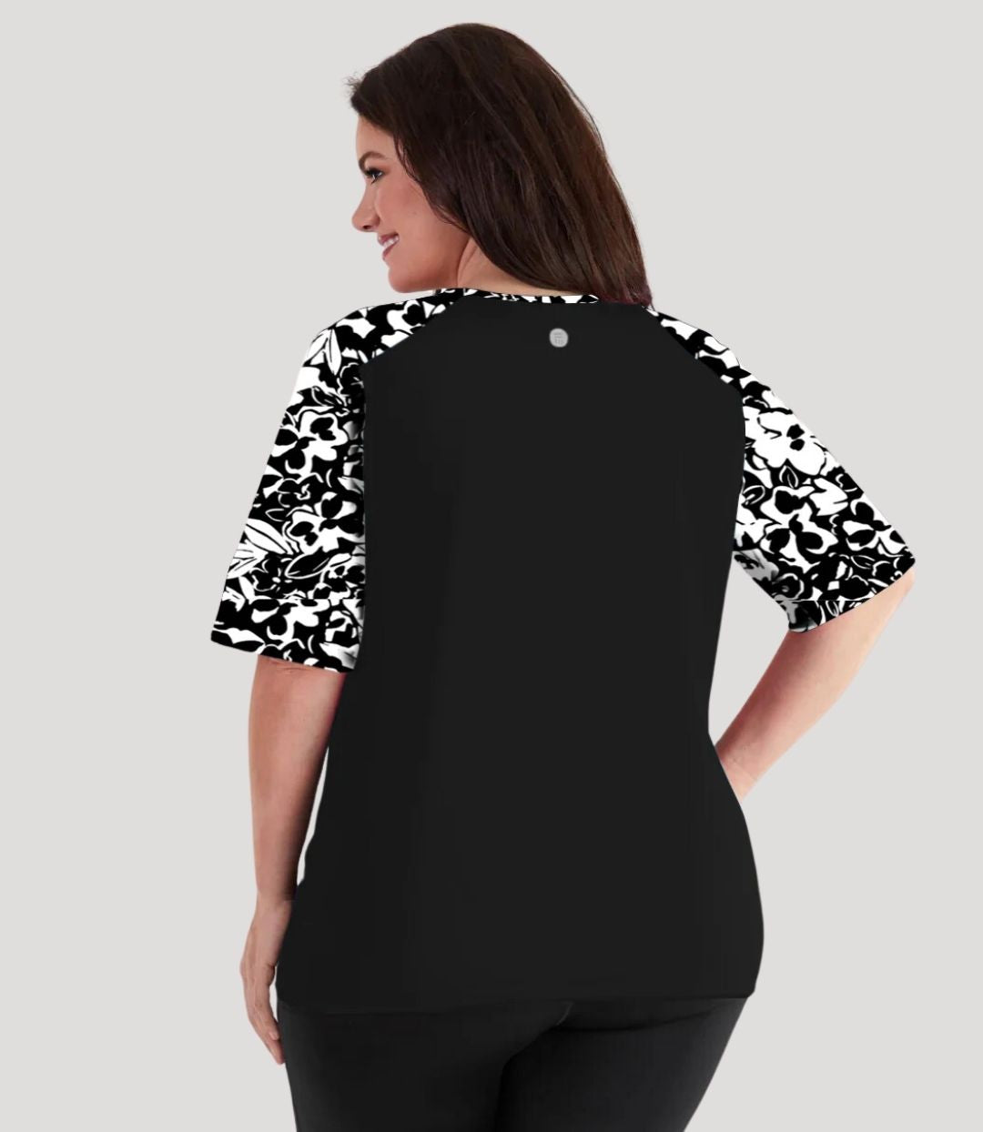 Plus size woman, facing back wearing JunoActive QuikEnergy Lite Swim and Beach short Sleeve Top Hibiscus Black. Solid Black on the center and Hibiscus print on the sleeves and upper shoulders. The short sleeves end at the elbow.