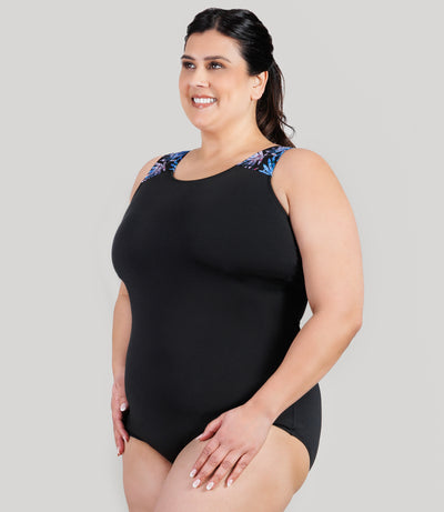 Plus size model, facing side, wearing JunoActive's AquaSport Crossback Plus Size Tanksuit in Black and Jewel Palmetto print.