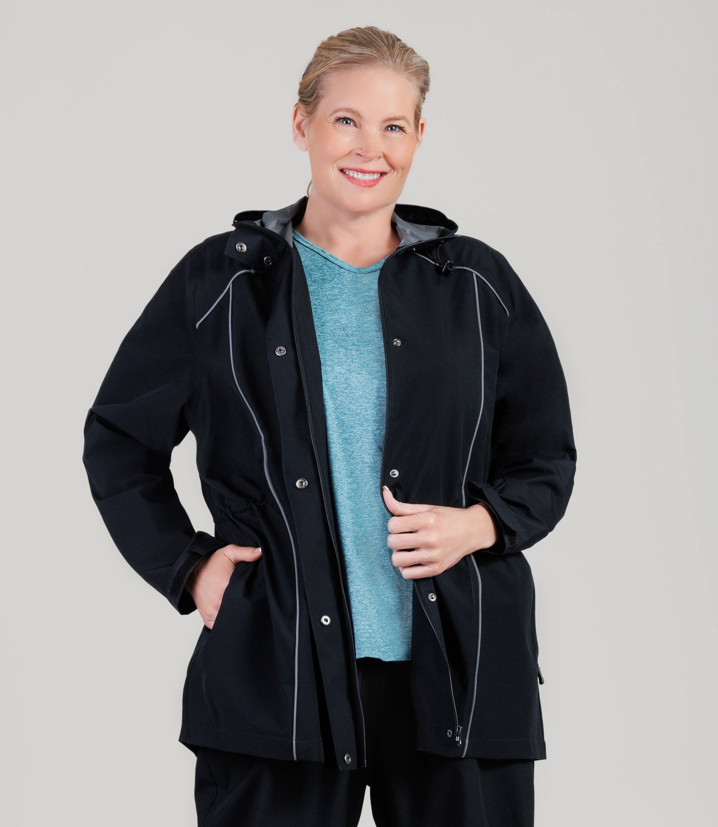 Plus size model, wearing JunoActive's Adventure Wind and Rain Jacket in color black. Jacket is unzipped with her right hand in the jacket pocket and left hand holding onto zipper of jacket.