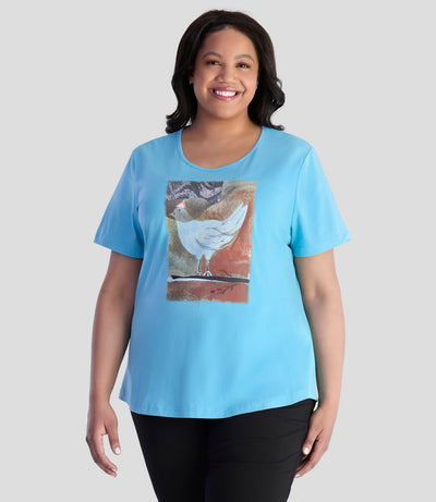 Model is facing forward with her arms by her side. She is wearing JunoActive's Designer Graphic scoop neck top with bird print on color mountain lake blue. 