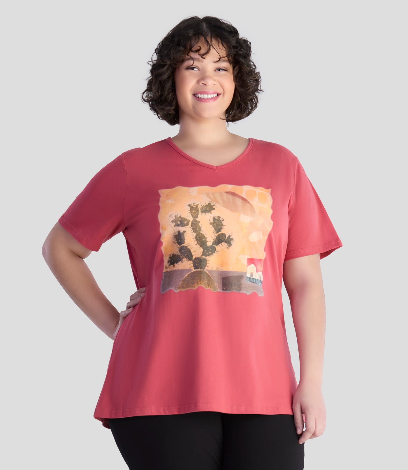 Model is wearing JunoActive's designer graphic v-neck top in rosy red. Printed is a Sedona scene with cactus, a house in the background and lovely warm colors with unique texture. Models right hand is on her hip and left arm is by her side. 
