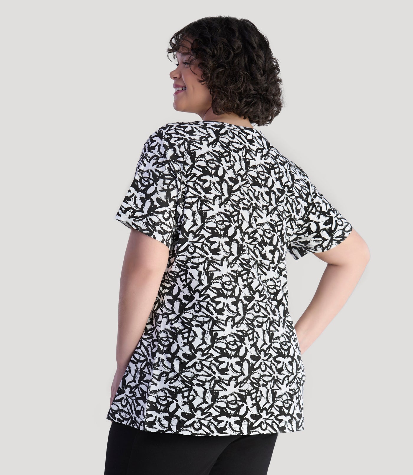 Model, facing back, wearing JunoActive's Junonia Lifestyle printed short sleeve neck top in color black and white wildflower print.