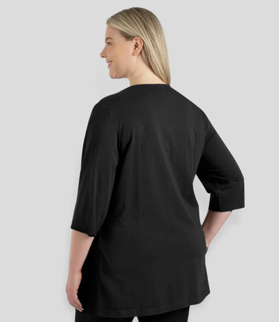Plus size woman, facing back, wearing JunoActive’s Stretch Naturals Lite 3/4 Sleeve V-neck Tunic, color black. Left arm by side & right hand on hip.