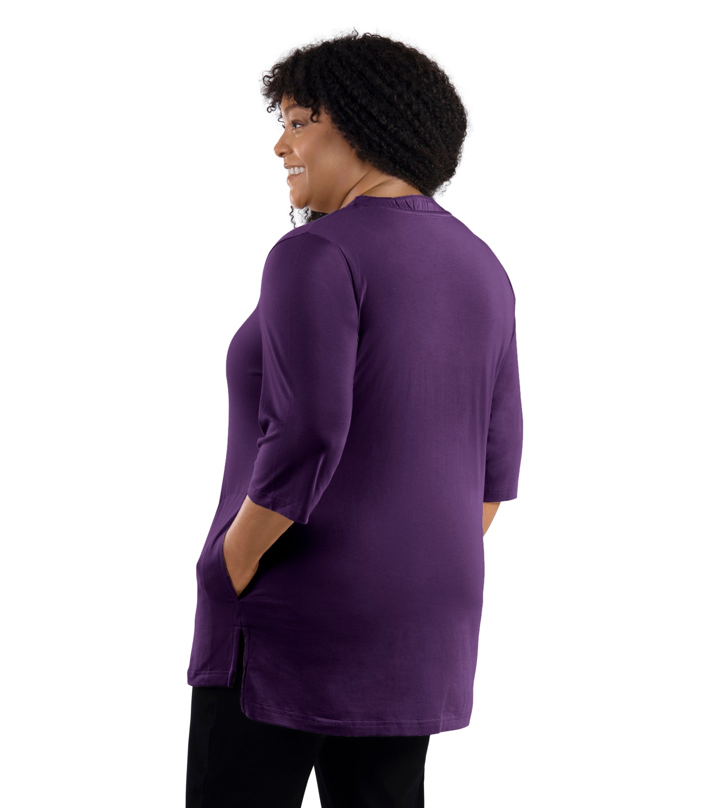 JunoActive Model wearing Stretch Naturals Lite 3/4 Sleeve Button Henley, facing back, one hand in pocket of shirt and other by her side. Color deep plum. 