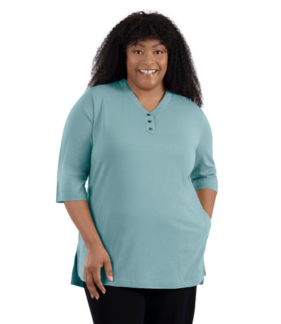 JunoActive Model wearing Stretch Naturals Lite 3/4 Sleeve Button Henley, facing front, one hand in pocket of shirt and other by her side. Color calm jade.