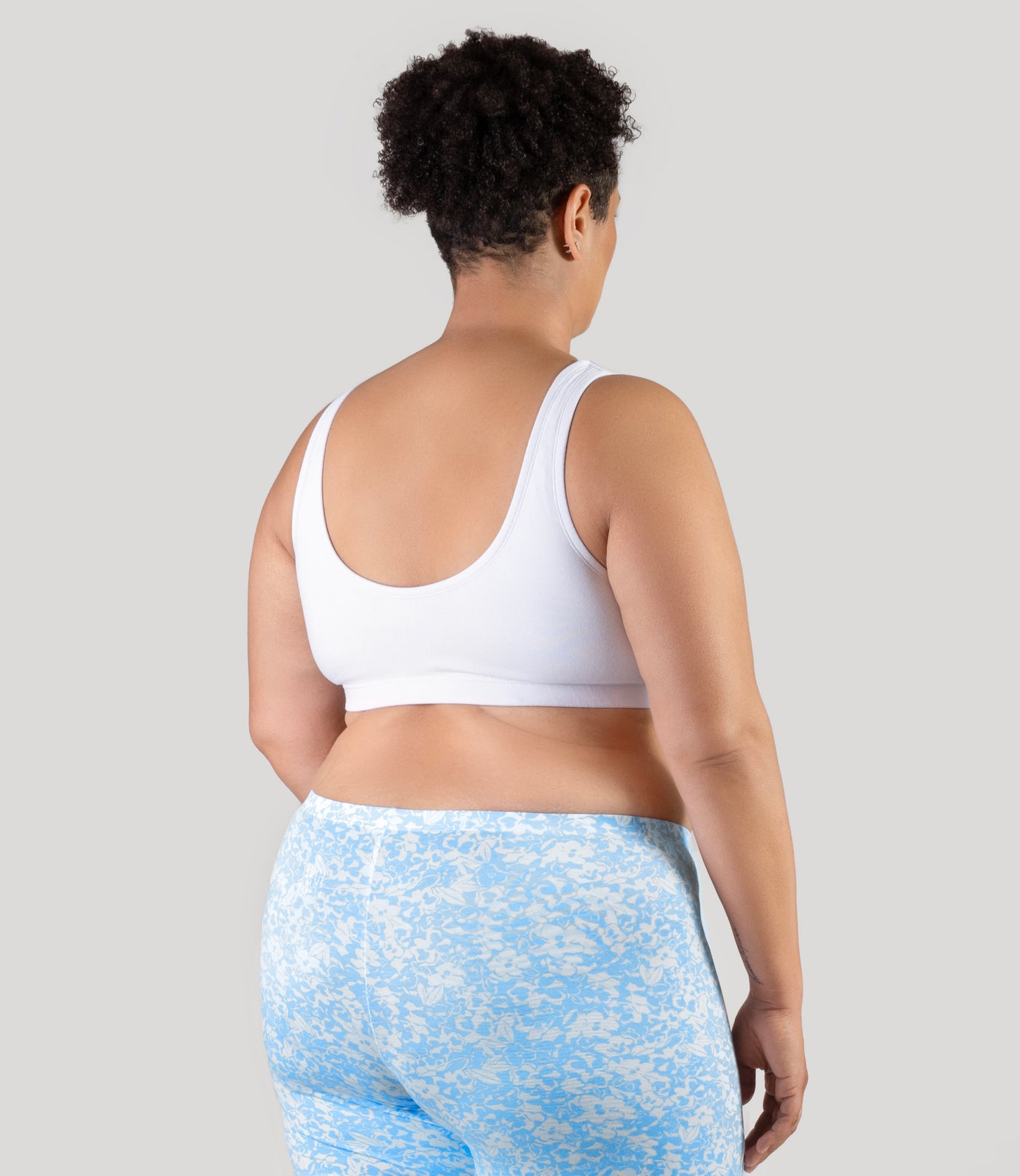 Plus size woman, facing to the back, both hands by side, wearing JunoActive Stretch Naturals Shirred plus size Bra Top. plus size sports Bra is gathered in middle front and has a scoop neck and back. Straps are wide and bra color is white.