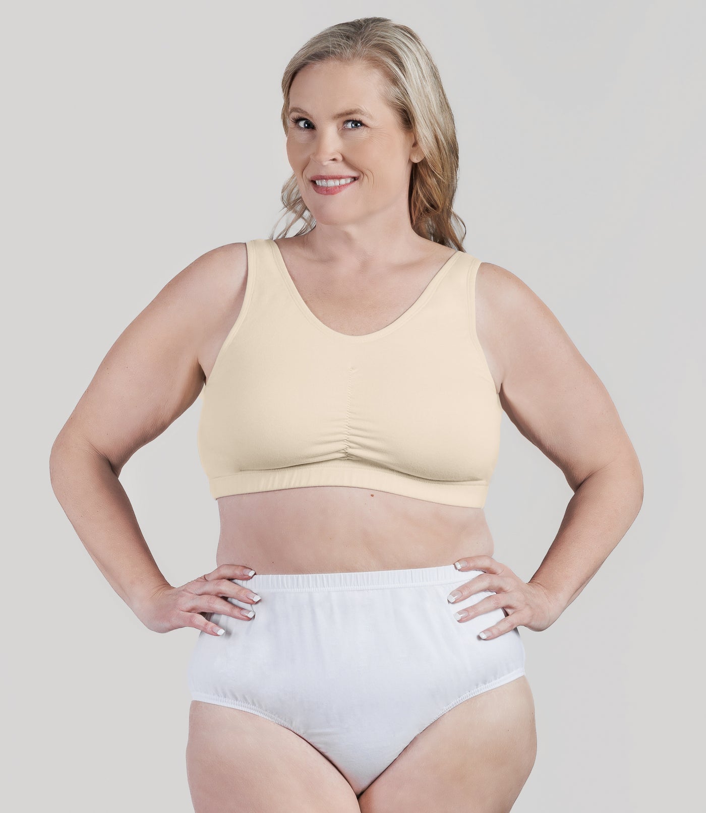 Plus size woman, facing front, wearing JunoActive Stretch Naturals Shirred Bra Top. Bra is gathered in middle and is a scoop top neck. Straps are wide and bra color is sandy.