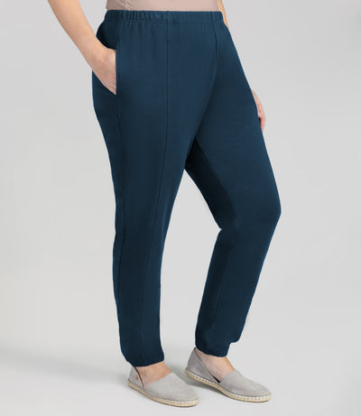 Plus size woman facing forward wearing JunoActive Stretch Naturals plus size jogger pant bottom. Her right hand is in pocket and left arm hanging by side. The pant is shirred at top with elastic band and loosely fitted. The hem at bottom is gathered with elastic at ankle. Color is indigo.