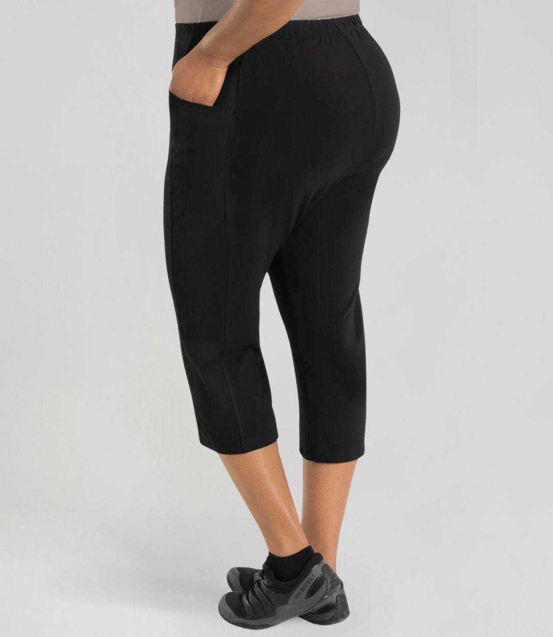 Plus size woman facing to the side and back with left hand in pocket wearing JunoActive Stretch Naturals side pocket plus size capris. The hem falls mid-calf and is form fitting without being skin tight. Color is black.