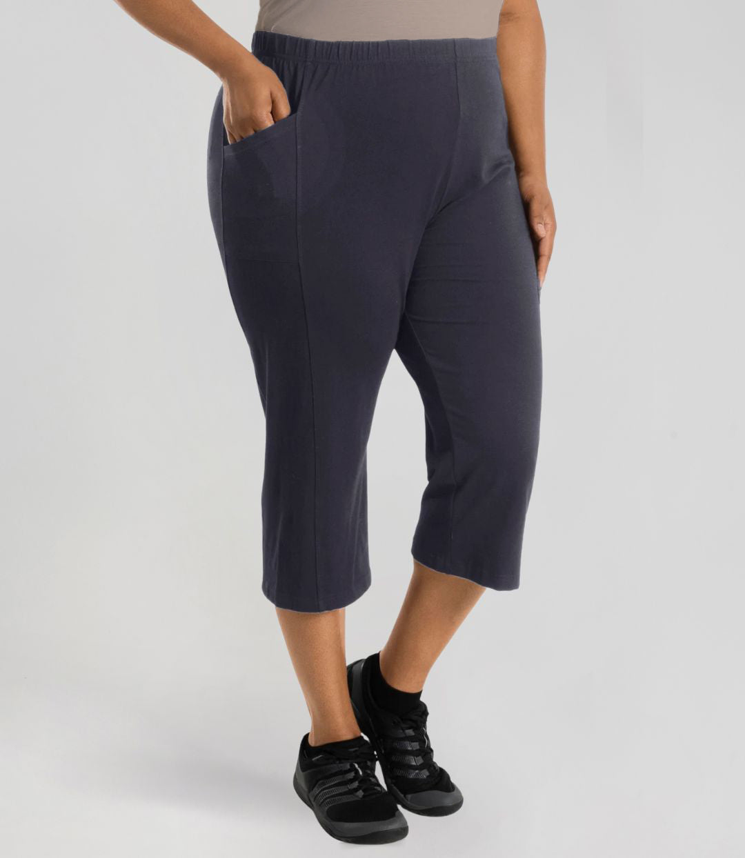 Plus size woman is facing at a side angle, right hand in pocket, left arm and hand to side, wearing JunoActive Stretch Naturals side pocket plus size capri pant bottom. The hemline comes to mid-calf and hugs the body without being skin tight. Color is oak gray.