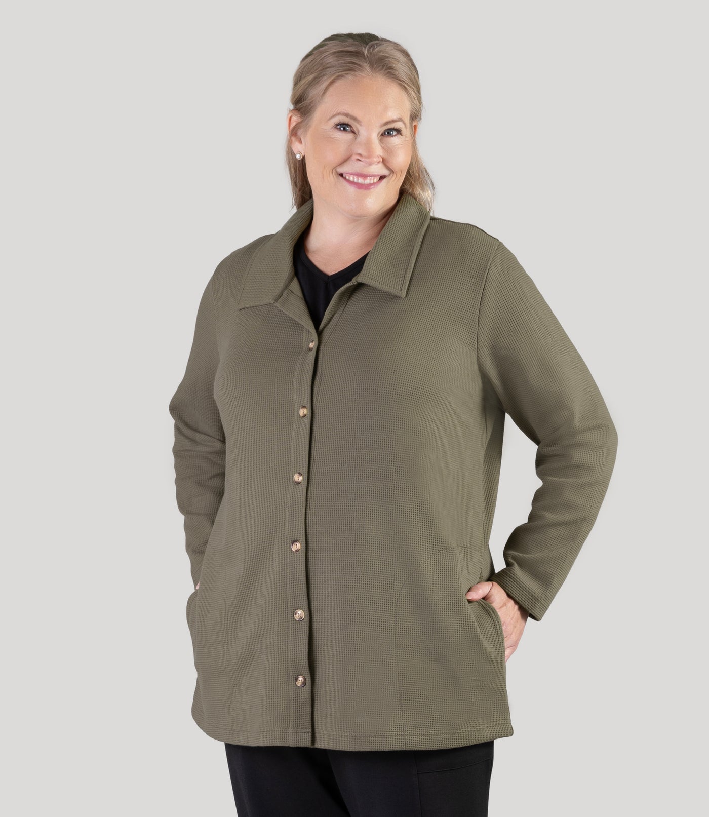 Plus size model, facing front, wearing JunoActive's JunoWaffle Cotton Long Sleeve Button top with her hands in her pockets of top. Color is in olive.